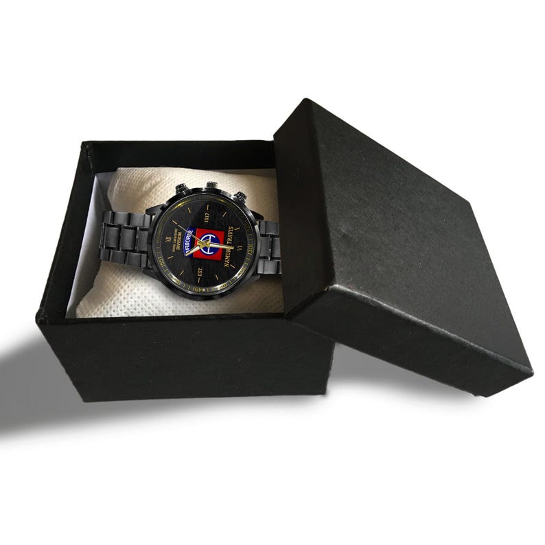 May include: A black wristwatch with a matching metal band is displayed inside an open black box. The watch face features a design with red, white, and blue colors. The watch is presented on a white, textured fabric.