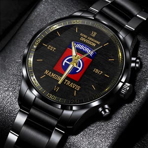 May include: Black wristwatch with a round face featuring gold accents and a black band. The watch face displays the text "82ND AIRBORNE DIVISION" and "NAMON TRAVIS" along with a red, white, and blue emblem. The watch has a metallic finish.