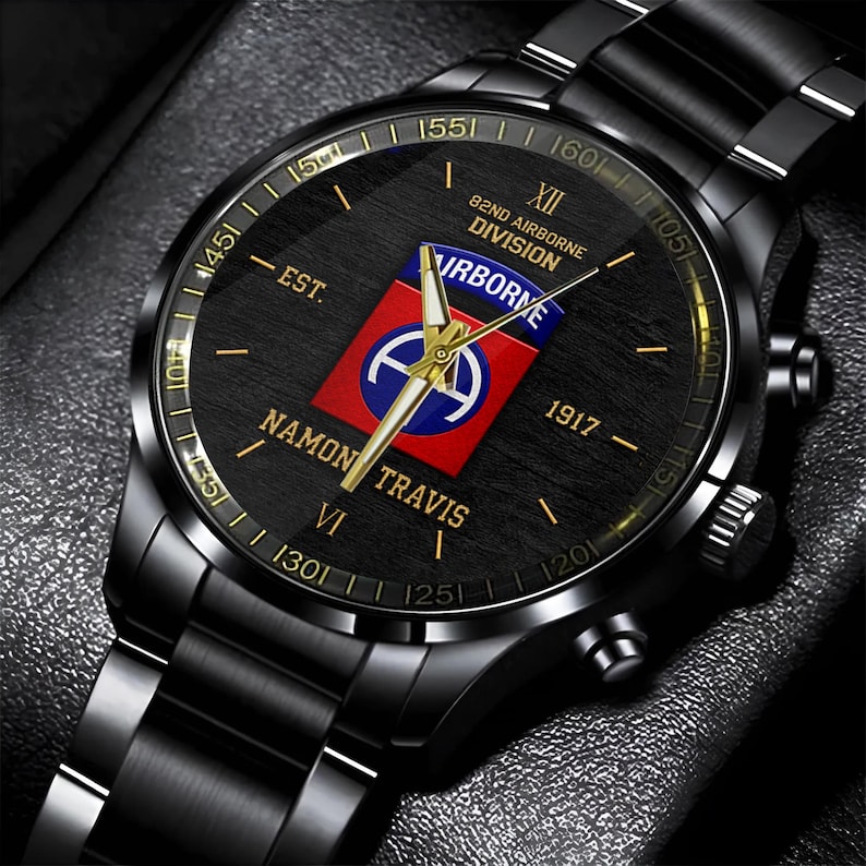 May include: Black wristwatch with a round face featuring gold accents and a black band. The watch face displays the text "82ND AIRBORNE DIVISION" and "NAMON TRAVIS" along with a red, white, and blue emblem. The watch has a metallic finish.