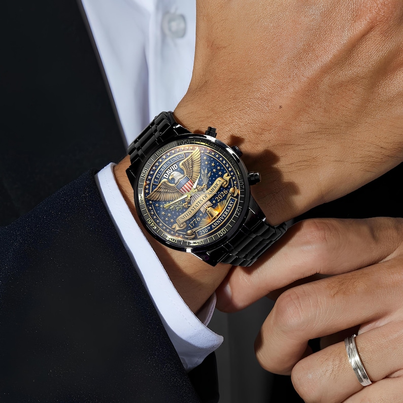 May include: Black wristwatch with a detailed face featuring an eagle emblem, stars, and the text "United States of America 1776-2026". The watch has a black metal band and is worn on a wrist. A silver ring is visible on the finger.