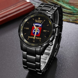 May include: Black wristwatch with a black metal band. The watch face features the text "82ND AIRBORNE DIVISION" and "NAMON TRAVIS". The watch face also has a red, white, and blue emblem. The watch has gold-colored accents.