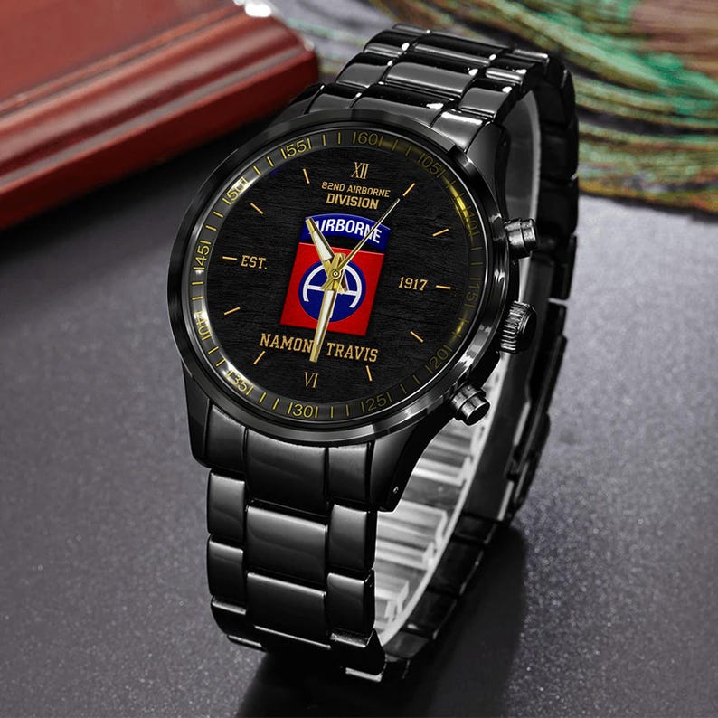 May include: Black wristwatch with a black metal band. The watch face features the text "82ND AIRBORNE DIVISION" and "NAMON TRAVIS". The watch face also has a red, white, and blue emblem. The watch has gold-colored accents.