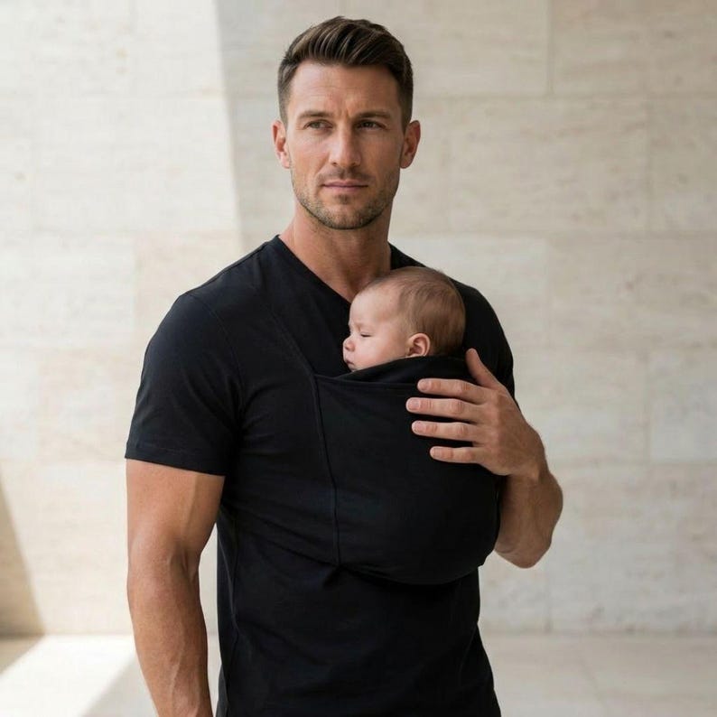 May include: A black baby carrier holds a sleeping infant. The carrier is worn by a person wearing a black V-neck t-shirt. The background is a neutral, light-colored wall.