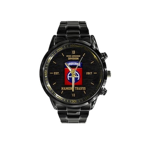 May include: Black wristwatch with a round face. The watch face features the text "B2ND AIRBORNE DIVISION" and "NAMON TRAVIS". The watch has a black metal band and a black bezel. The watch face has a red, white, and blue emblem.