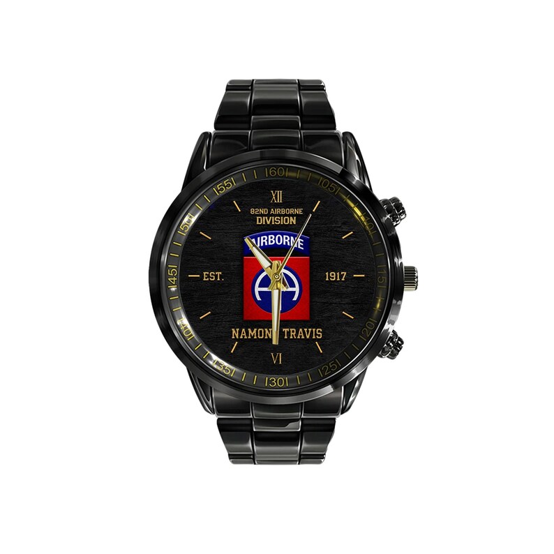 May include: Black wristwatch with a round face. The watch face features the text "B2ND AIRBORNE DIVISION" and "NAMON TRAVIS". The watch has a black metal band and a black bezel. The watch face has a red, white, and blue emblem.