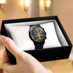 May include: A black wristwatch with a detailed face featuring an eagle emblem and the text "UNITED STATES OF AMERICA 1776 - 2026 250th ANNIVERSARY". The watch is presented in a black gift box with a white cushion.