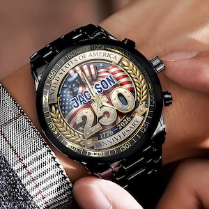 May include: Black wristwatch with a round face featuring the American flag and the text "JACKSON 250 ANNIVERSARY 1776-2026". The watch has a black metal band and a gold-colored bezel with the words "UNITED STATES OF AMERICA".