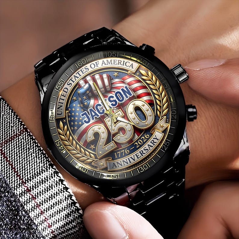 May include: Black wristwatch with a round face featuring the American flag and the text "JACKSON 250 ANNIVERSARY 1776-2026". The watch has a black metal band and a gold-colored bezel with the words "UNITED STATES OF AMERICA".