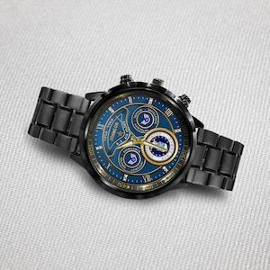 May include: A black stainless steel wristwatch with a blue dial featuring gold-tone hands and markers. The dial displays the text Johnson, 1956-1969, U.S. Air Force, Excellence, and Integrity, along with three circular sub-dials and Roman numerals.