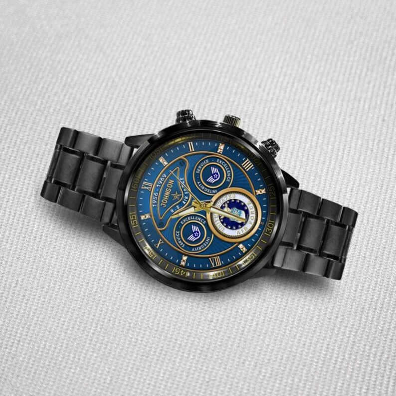 May include: A black stainless steel wristwatch with a blue dial featuring gold-tone hands and markers. The dial displays the text Johnson, 1956-1969, U.S. Air Force, Excellence, and Integrity, along with three circular sub-dials and Roman numerals.