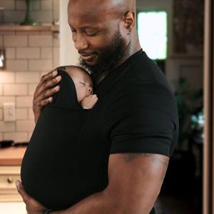 May include: A black baby carrier holds a sleeping infant. The carrier is worn by a person wearing a black shirt. The baby is nestled in the carrier, with a hood over its head. The background is a softly lit interior.
