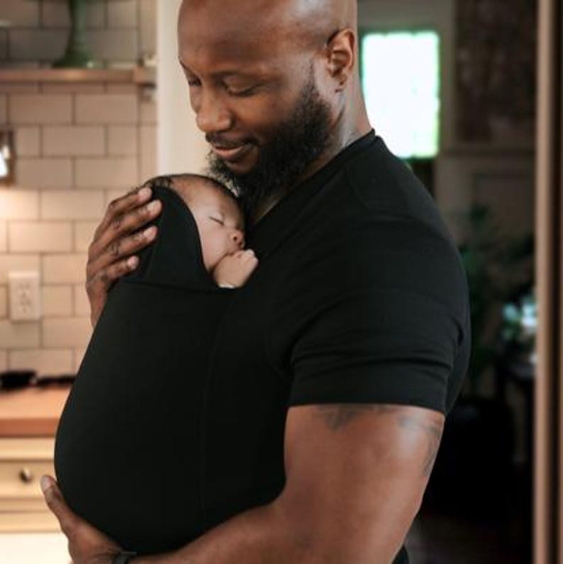 May include: A black baby carrier holds a sleeping infant. The carrier is worn by a person wearing a black shirt. The baby is nestled in the carrier, with a hood over its head. The background is a softly lit interior.