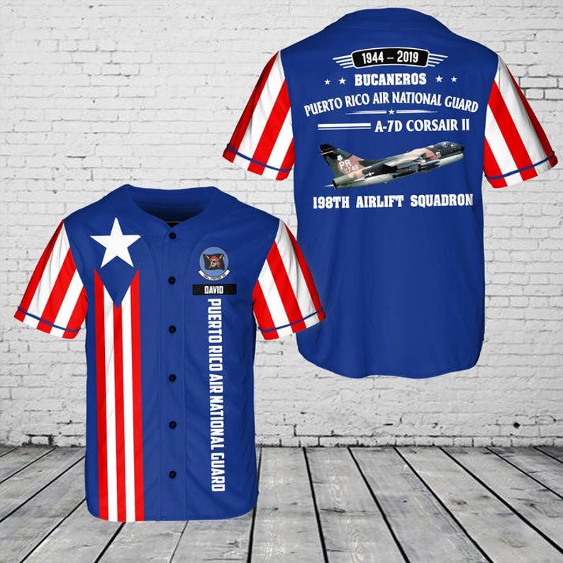 May include: A blue baseball jersey with red and white striped sleeves. The front features a white star and the Puerto Rican flag design. The back displays text: "1944-2019 BUCANEROS PUERTO RICO AIR NATIONAL GUARD A-7D CORSAIR II 198TH AIRLIFT SQUADRON."