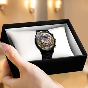 May include: A black wristwatch with a gold-colored face featuring the text "JACKSON 250 ANNIVERSARY 1776-2026" and an American flag design. The watch is displayed in a black box on a white pillow.