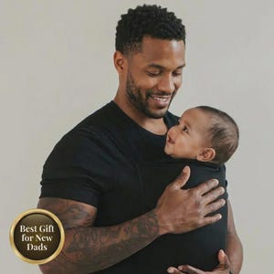 May include: A smiling person wearing a black t-shirt holds a baby in a black baby carrier. The baby is looking up at the person. A gold emblem in the lower left corner reads "Best Gift for New Dads."