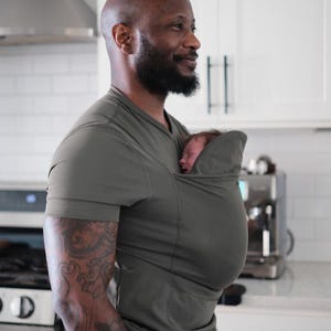 May include: Olive green baby carrier shirt. The shirt has a pouch to hold a baby. The person wearing the shirt has a bald head and a beard. The person is smiling and looking to the side. The background is a kitchen.