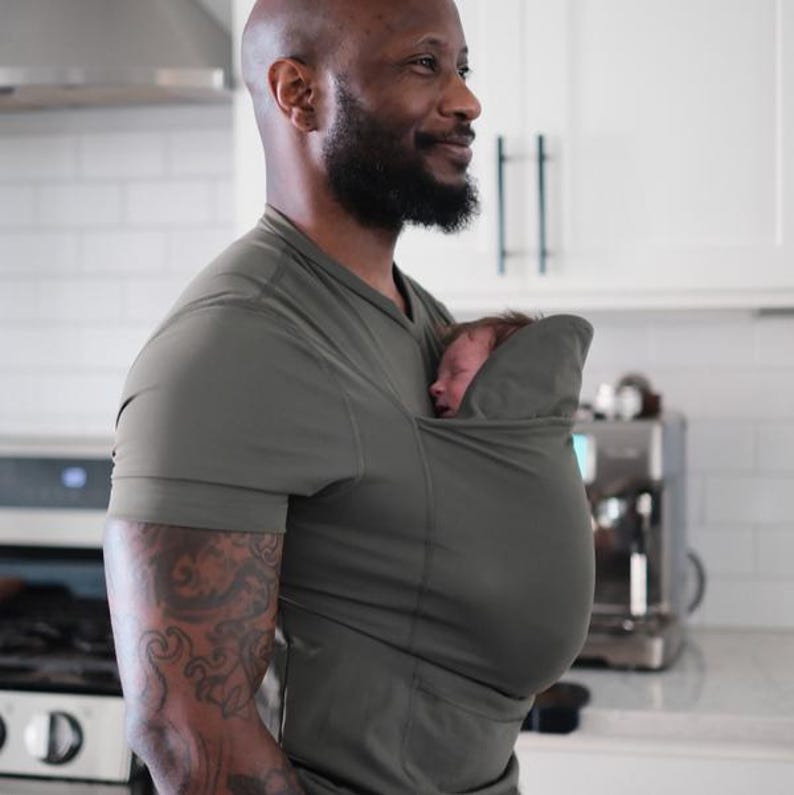 May include: Olive green baby carrier shirt. The shirt has a pouch to hold a baby. The person wearing the shirt has a bald head and a beard. The person is smiling and looking to the side. The background is a kitchen.
