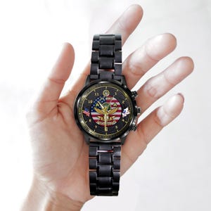 May include: A black metal wristwatch with a round face featuring a U.S. Army emblem, an American flag background, and the word VETERAN printed in gold. The watch has gold-coloured hands, numerical hour markers, and a black link bracelet strap.