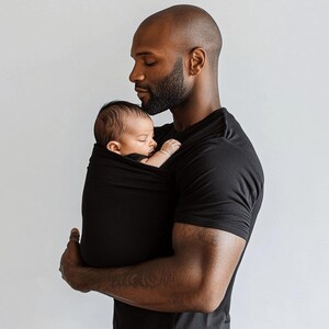 May include: A black baby wrap carrier cradles a sleeping infant. The wrap is held by a person wearing a black t-shirt. The background is a plain white wall.