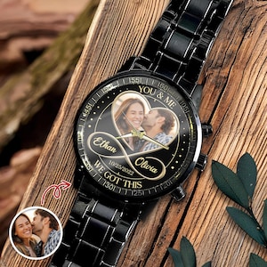 May include: A black metal wristwatch with a circular face featuring a heart-shaped photo of a couple, the names Ethan and Olivia, an infinity symbol, the date 10/25/2022, and the text YOU & ME and WE GOT THIS printed on the black dial.