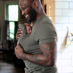 May include: An olive green baby carrier holds a sleeping infant. The carrier is integrated into a short-sleeved shirt. The person wearing the shirt has a beard and tattoos on their arm. The background is a domestic interior.