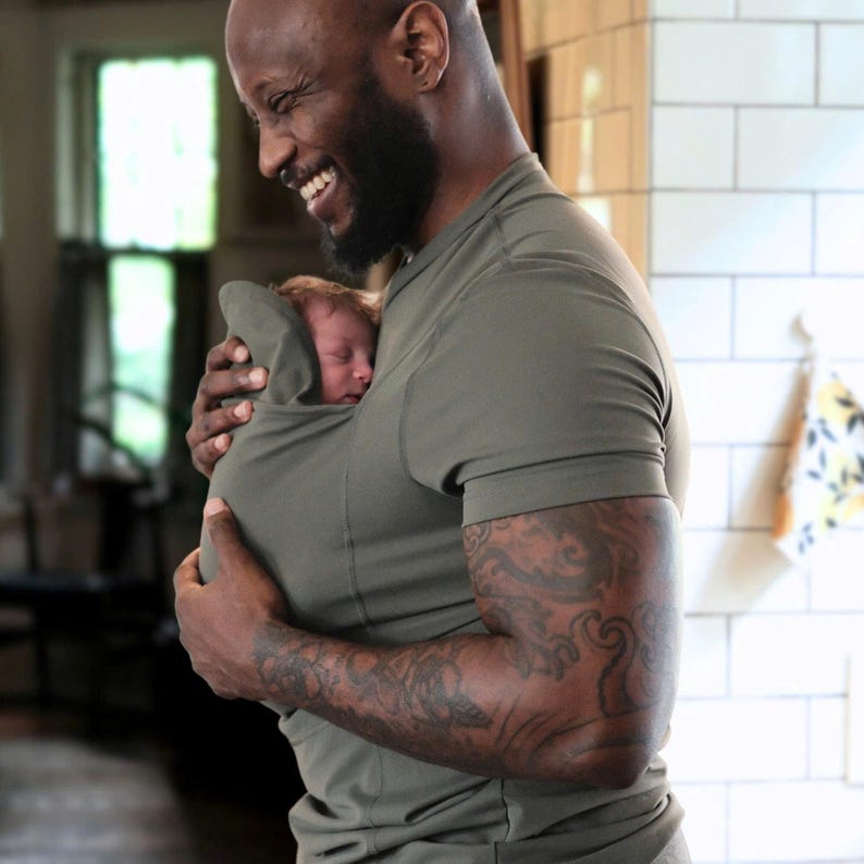 May include: An olive green baby carrier holds a sleeping infant. The carrier is integrated into a short-sleeved shirt. The person wearing the shirt has a beard and tattoos on their arm. The background is a domestic interior.