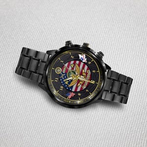 May include: A black metal wristwatch with a round face featuring a U.S. Army emblem, a gold eagle graphic, and a red, white, and blue striped background. The dial includes the text U.S. ARMY and VETERAN, with gold hands and numerical hour markers.