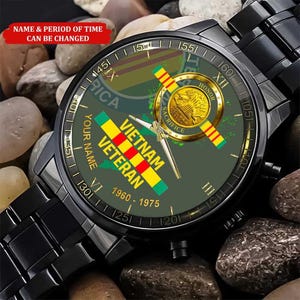 Puede incluir: Reloj de pulsera negro con esfera verde con un dise&ntilde;o de Veterano de Vietnam. La esfera incluye el texto "Vietnam Veteran" y "1960-1975", junto con un emblema dorado y una correa negra. El reloj se muestra sobre un lecho de peque&ntilde;as piedras.