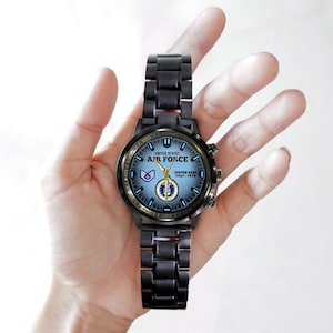 May include: A black metal wristwatch with a light blue dial featuring the text United States Air Force, a military emblem, and the custom text Custom Name 1967-1978. The watch has a link bracelet, gold-coloured hands, and a circular case.