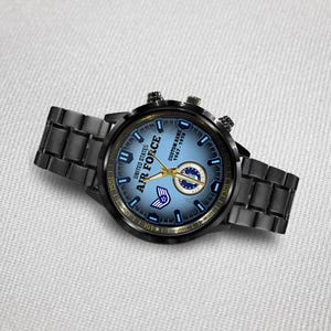 May include: A black metal wristwatch with a blue gradient dial featuring the text UNITED STATES AIR FORCE, CUSTOM NAME, and 1967-1978. The watch includes gold-toned hands, a circular emblem, and a small wing icon on the face with a link bracelet.