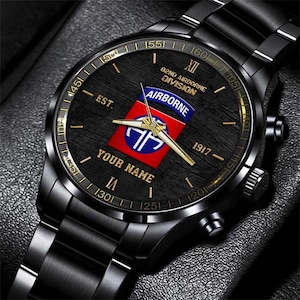 May include: Black wristwatch with a black face and band. The watch face features gold numerals and hands, with the text "YOUR NAME" and a red, white, and blue emblem that reads "AIRBORNE". The watch also has the text "82ND AIRBORNE DIVISION" and "EST. 1917".