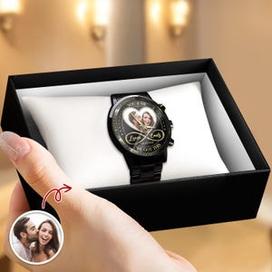 May include: A black wristwatch with a custom photo dial featuring a couple, the text "YOU & ME", "Lucas", "Emily", "08/18/2020", and "WE GOT THIS" inside a heart and infinity symbol, presented on a white cushion inside a black gift box.