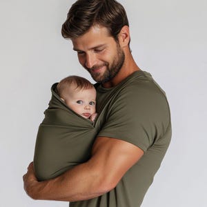 May include: An olive green baby carrier holds a baby. The baby is wrapped in the same olive green fabric. The person holding the baby is wearing an olive green t-shirt. The background is a plain white.
