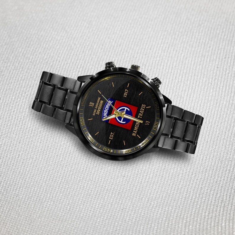 May include: Black wristwatch with a round face and a matching black metal band. The watch face features a military emblem with the text "82nd Airborne Division" and "Namon Travis". The watch has gold accents and a textured black background.