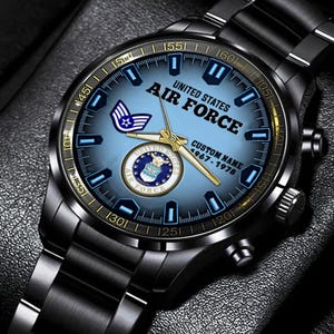 May include: A black stainless steel wristwatch with a light blue dial featuring the United States Air Force emblem, the text UNITED STATES AIR FORCE, CUSTOM NAME, and 1967-1978. The watch has gold-toned hands and a black metal link bracelet.