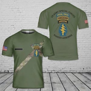 May include: Olive green t-shirt featuring the US Army Special Forces insignia. The front displays a camouflage design and the American flag. The back of the shirt has the text "US Army Special Forces" and "Ranger Airborne" with a blue and yellow emblem.