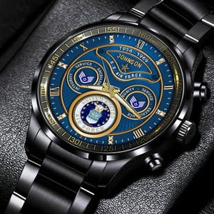 May include: A black metal wristwatch with a blue dial featuring the United States Air Force emblem, the text Johnson 1956-1969, and gold-toned hands. The watch face includes three sub-dials, gold Roman numeral hour markers, and small crystal accents.