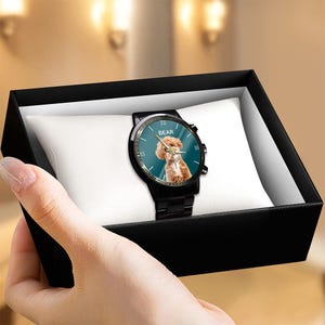 May include: A black wristwatch with a metal link band, featuring a teal watch face with a graphic of a brown dog and the word BEAR printed in white. The watch includes gold-toned hands and Roman numeral markers, presented on a white cushion.