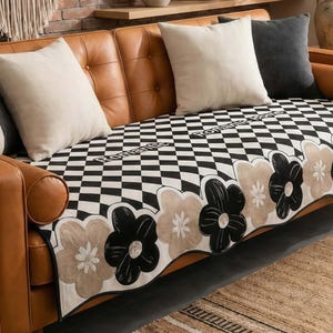 May include: A brown leather sofa with cream and gray throw pillows. A black and white checkered sofa cover with a floral border is draped over the sofa. The sofa is in a living room setting.
