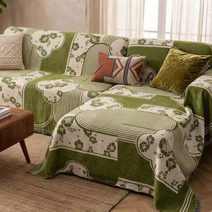 May include: A green and cream-colored sofa cover with a floral and geometric pattern. The cover is draped over a sectional sofa, with several decorative pillows in various colors and textures. A wooden coffee table and a woven rug are in the foreground.