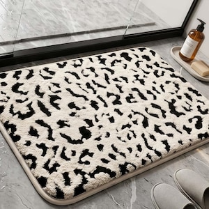 May include: A rectangular bath mat featuring a high-pile, textured surface with a black and cream leopard print pattern. The mat has rounded corners and a solid cream-colored border, designed for placement on a smooth, tiled bathroom floor.