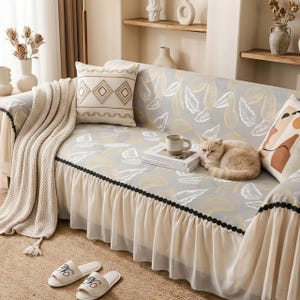 Può includere: Un divano con una fodera grigia a motivi di foglie, una gonna color crema e cuscini decorativi. Una coperta beige e un paio di pantofole bianche con iniziali ricamate nere sono sul pavimento. Un gatto riposa sul divano.