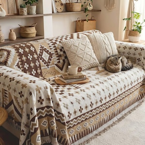 Puede incluir: Un sofá cubierto con una manta estampada en crema y marrón. Cojines decorativos en crema y beige descansan sobre el sofá. Un gato está acurrucado en el sofá. Una bandeja de madera con libros y una taza está en el sofá.