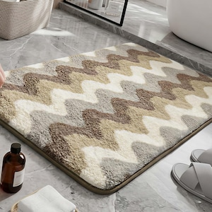 May include: A rectangular bath mat featuring a textured, shaggy pile with a repeating chevron pattern in shades of cream, beige, light grey, and dark brown. The rug has rounded corners and a solid-coloured border, providing a soft surface.