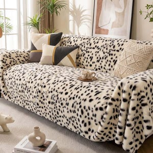 Könnte beinhalten: Ein Sofa, das mit einer weiß-schwarz gefleckten Decke bedeckt ist. Dekorative Kissen mit geometrischen Mustern und ein Makramee-Kissen liegen auf dem Sofa. Eine Tasse Kaffee steht auf einer kleinen Untertasse auf dem Sofa.