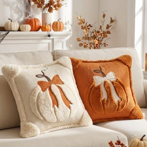 Puede incluir: Dos cojines cuadrados con diseños de calabaza texturizados. El cojín izquierdo es crema con una calabaza y un lazo naranja, mientras que el derecho es naranja con una calabaza y un lazo crema. Ambos tienen aplicaciones de felpa suave.