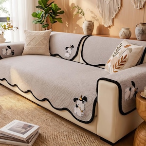 以下が含まれることがあります： 波状の黒いトリムと漫画の犬のアクセントが付いた、明るい灰色のソファカバー。 カバーは質感があり、ベージュのニットクッションと花柄のクッションが特徴です。 ソファはクリーム色のベースです。