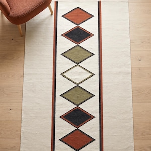 May include: A rectangular area rug featuring a cream-colored base with a central vertical band containing seven diamond shapes in alternating colors of rust orange, black, and olive green, framed by thin black and rust orange vertical stripes.