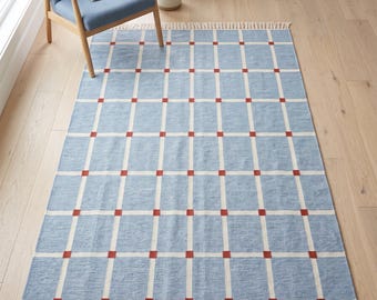 May include: A rectangular area rug featuring a light blue base with a white grid pattern and small red squares at each intersection. The rug has a woven texture and is finished with cream-colored fringe along the top and bottom edges.