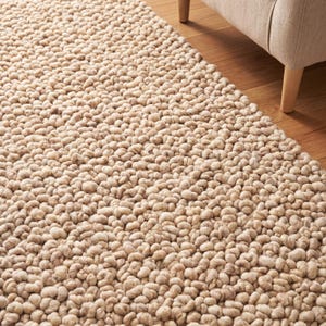 Puede incluir: Una alfombra beige con una pila densa y en bucle, con una superficie texturizada. El diseño de la alfombra presenta pequeños nudos redondeados, creando un patrón táctil. La alfombra está colocada sobre un suelo de madera, con parte de un sillón beige.